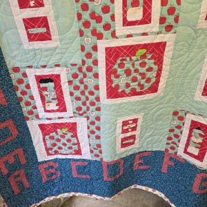 May include: A patchwork quilt featuring a light blue background with a repeating red apple pattern. The quilt includes white-bordered blocks displaying various apple-themed illustrations and red fabric letters spelling out the alphabet.