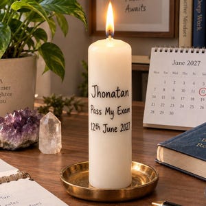 Puede incluir: Una vela de pilar blanca con una llama encendida se asienta sobre una bandeja redonda de metal dorado. La vela presenta texto impreso en negro que dice Jhonatan, Pass My Exam y 12th June 2027. La vela está colocada sobre una superficie de escritorio de madera.