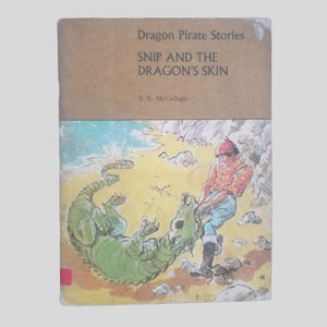 Peut inclure: Couverture de livre pour enfants vintage, "Dragon Pirate Stories: Snip and the Dragon's Skin" de S. K. McCullagh. L'illustration montre un dragon vert et une personne en chapeau rouge et chemise orange sur une plage.