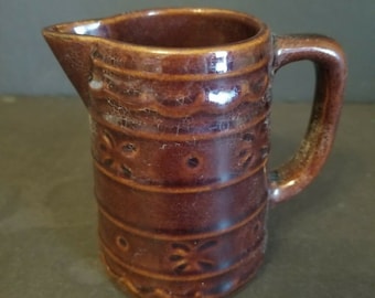 May include: A small, brown ceramic pitcher with a handle and a spout. The pitcher is decorated with horizontal bands and floral patterns. The glaze has a crackled appearance, adding to its vintage charm. This decorative pitcher is suitable for serving liquids.