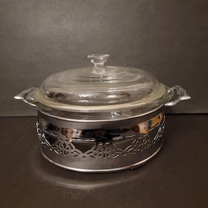 May include: A silver-toned glass casserole dish with a clear glass lid and a decorative metal base. The base has a delicate, intricate design.