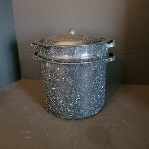 May include: A large, speckled black and white enamelware pot with a lid. The pot has two side handles and a lid with a small handle. The pot is designed for cooking and canning. The pot is sitting on a dark surface.