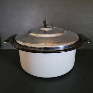 May include: A white enamel pot with a silver lid and black handles. The lid has a black finial. The pot is sitting on a black surface against a dark background. This vintage cookware is suitable for cooking.