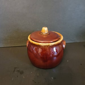 May include: A brown ceramic jar with a lid and two handles. The jar has a glazed finish and a light brown rim.