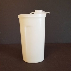 May include: A tall, off-white plastic tumbler with a matching lid and a flip-top opening. The cylindrical container stands upright against a dark background. The lid is attached to the top of the tumbler.