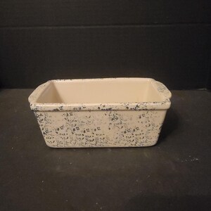 May include: A white ceramic loaf pan with a speckled blue glaze. The pan has a rectangular shape with rounded corners and two small handles on the sides.