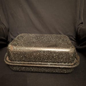 May include: A rectangular, speckled black roasting pan with a lid. The pan has a dark speckled pattern with white flecks. The pan is made of metal and has a rim around the top and bottom. The pan is designed for cooking.