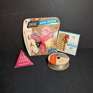 May include: A collection of crafting supplies including a Pixie Bow Maker with a pink plastic tool, a Chadwick Bowmaker Kit in a rectangular box, a triangular box of 100 Pixie Bow Pins, and a spool of silver Lace-Sheen ribbon measuring 20 feet.
