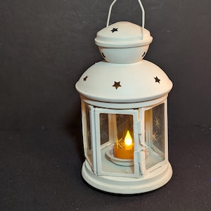 May include: A white metal lantern featuring a hinged door and cut-out star shapes on the top dome. The glass panels are etched with star designs, and a small, battery-operated yellow LED tea light candle sits inside the central chamber.