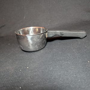 May include: A stainless steel measuring cup with a black plastic handle. The cup is round with a slightly flared rim. The handle is attached to the side of the cup and is rectangular in shape. The cup is sitting on a black surface.