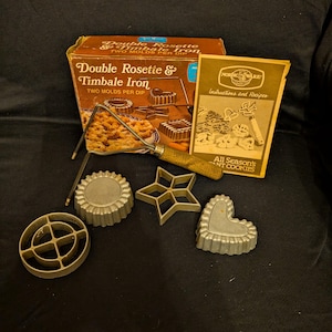 May include: A vintage Double Rosette & Timbale Iron set with a box, instructions, and various metal cookie molds. The molds include a circle, star, heart, and other shapes. The box has text that reads "Double Rosette & Timbale Iron".
