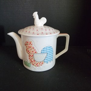 May include: White ceramic teapot with a rooster design. The teapot has a lid with a rooster finial and a patchwork design of red, blue, and green.