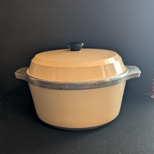 May include: A cream-colored Dutch oven with a matching lid and a small black knob handle. The pot features a metallic silver rim and two integrated side handles. The exterior has a smooth finish, and the overall design is simple and functional.