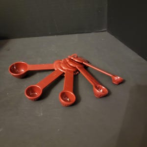 May include: A set of six red measuring spoons of varying sizes, connected by a ring. The spoons are made of a solid, smooth material and are arranged in a fan shape. The background is a dark gray surface.