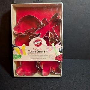 May include: A boxed set of red metal bug-shaped cookie cutters. The set includes six cutters in various shapes, including a butterfly, dragonfly, and ladybug. The packaging features the text "Bug Buddies Cookie Cutter Set".