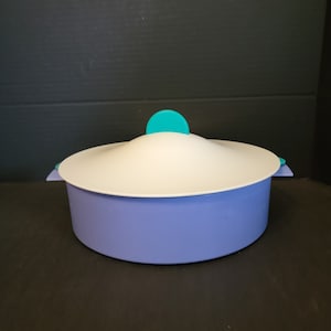 May include: A round, light purple storage container with a smooth, matte finish. It features a white, slightly domed lid with a bright teal circular handle on top and matching teal side tabs for securing the lid to the base of the container.