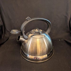 May include: Stainless steel stovetop kettle with a black handle and spout. The kettle has a shiny, reflective surface and a black base. The spout has a small, round opening. The handle is arched and attached to the top of the kettle.