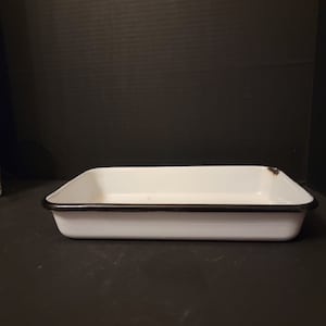 May include: A rectangular white enamelware baking dish with a black rim. The dish is empty and sits on a dark surface. The enamelware has a slightly distressed appearance.