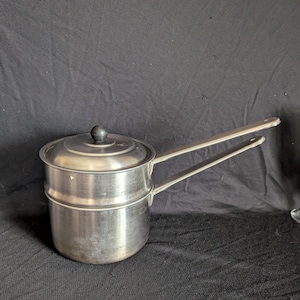 May include: A stainless steel double boiler with a lid and a black knob. The pot has two long handles extending from the sides. The background is a dark fabric.