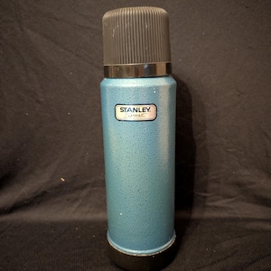 May include: A teal Stanley Sport thermos with a black cap and base. The thermos has a textured finish and a small rectangular label with the brand name. The overall design is cylindrical and classic.