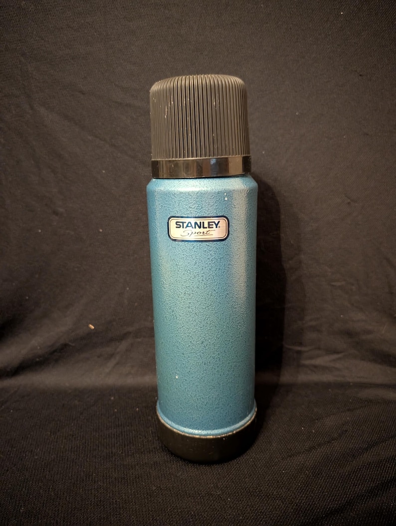 May include: A teal Stanley Sport thermos with a black cap and base. The thermos has a textured finish and a small rectangular label with the brand name. The overall design is cylindrical and classic.