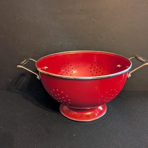 May include: A red colander with a silver rim and handles. The kitchen utensil has a round bowl shape with small holes for draining. It sits on a small, round base.