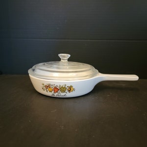 May include: White ceramic casserole dish with a glass lid. The dish has a floral design with a red pepper, green onion, and other vegetables. The text "Le Persil" is printed on the dish.