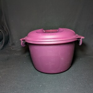May include: A deep purple Tupperware microwave container with a matching lid. The lid has a handle and two side clasps. The container is made of durable plastic and is designed for cooking and storing food.