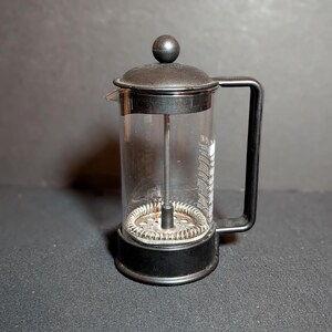 May include: A clear glass French press coffee maker featuring a black plastic lid with a spherical knob, a black plastic handle, and a black plastic base. A metal plunger mechanism with a coiled spring filter is visible inside the glass cylinder.