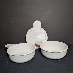 May include: Three white Corning Ware bowls with handles and a white lid with the text "Corning Ware" and other text. The bowls are round and have a simple, classic design. The lid is round with a handle.