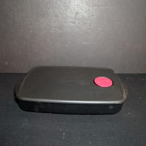May include: A black rectangular food storage container with a pink circular button on the lid. The container is made of a matte black material and has rounded corners. The lid appears to be securely attached.