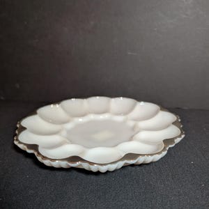 May include: White, scalloped-edge serving dish with a silver-colored trim. The dish has a central, shallow area surrounded by twelve rounded compartments. The dish is made of a glossy material and sits on a dark surface.