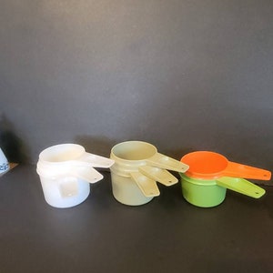 May include: Set of three nesting measuring cups. The largest cup is white, the middle cup is beige, and the smallest cup is green with an orange cup nested inside.