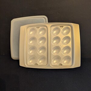 May include: A light beige plastic egg container with a clear lid. The container has two rows of oval-shaped indentations, each holding six eggs. The lid is rectangular and sits to the side of the container.