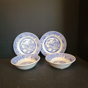 May include: Four-piece blue and white Willow pattern dish set. The set includes two plates and two bowls. The plates and bowls have a blue and white design with a detailed landscape scene. The dishes are arranged on a dark surface.