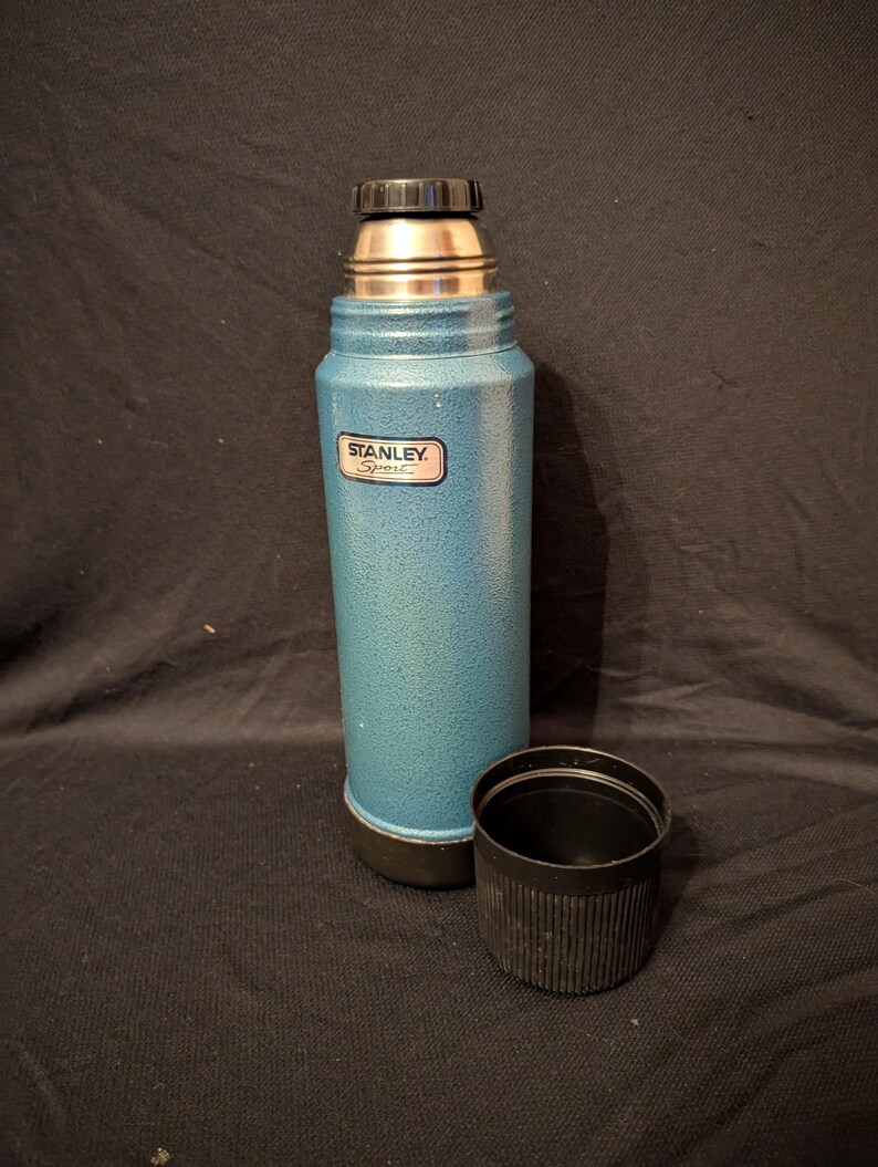 May include: A teal Stanley Sport thermos with a black cap and cup. The thermos has a silver top and a textured finish. The Stanley Sport logo is visible on the front. The cup is next to the thermos.