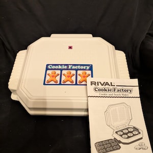 May include: A white Rival Cookie Factory cookie and snack maker with a gingerbread man sticker. The instruction manual is visible, with the text "Cookie Factory" and "Made in the USA".
