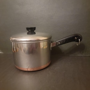 May include: A stainless steel saucepan with a lid and a black handle. The pot has a copper-colored bottom and a black knob on the lid. The handle has a loop for hanging. Kitchenware.