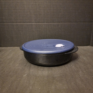 May include: A black plastic bowl with a dark blue lid. The lid has a small white vent and a textured surface. The bowl is sitting on a dark surface.
