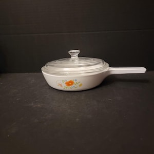 May include: White ceramic saucepan with a glass lid and a floral design. The flowers are orange, blue, and green.