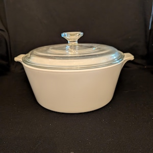 May include: A white casserole dish with a clear glass lid and a clear glass knob. The dish has two small handles on either side. The lid fits snugly on the dish. The dish is sitting on a black surface.