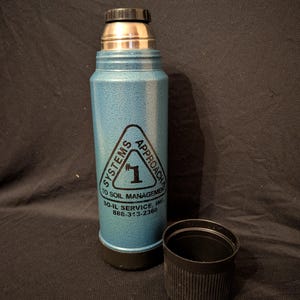 May include: A blue insulated thermos with a black lid and base, and a silver top. The thermos has a triangular logo with the text "SYSTEMS APPROACH TO SOIL MANAGEMENT" and the number "1". The black cap is next to the thermos.