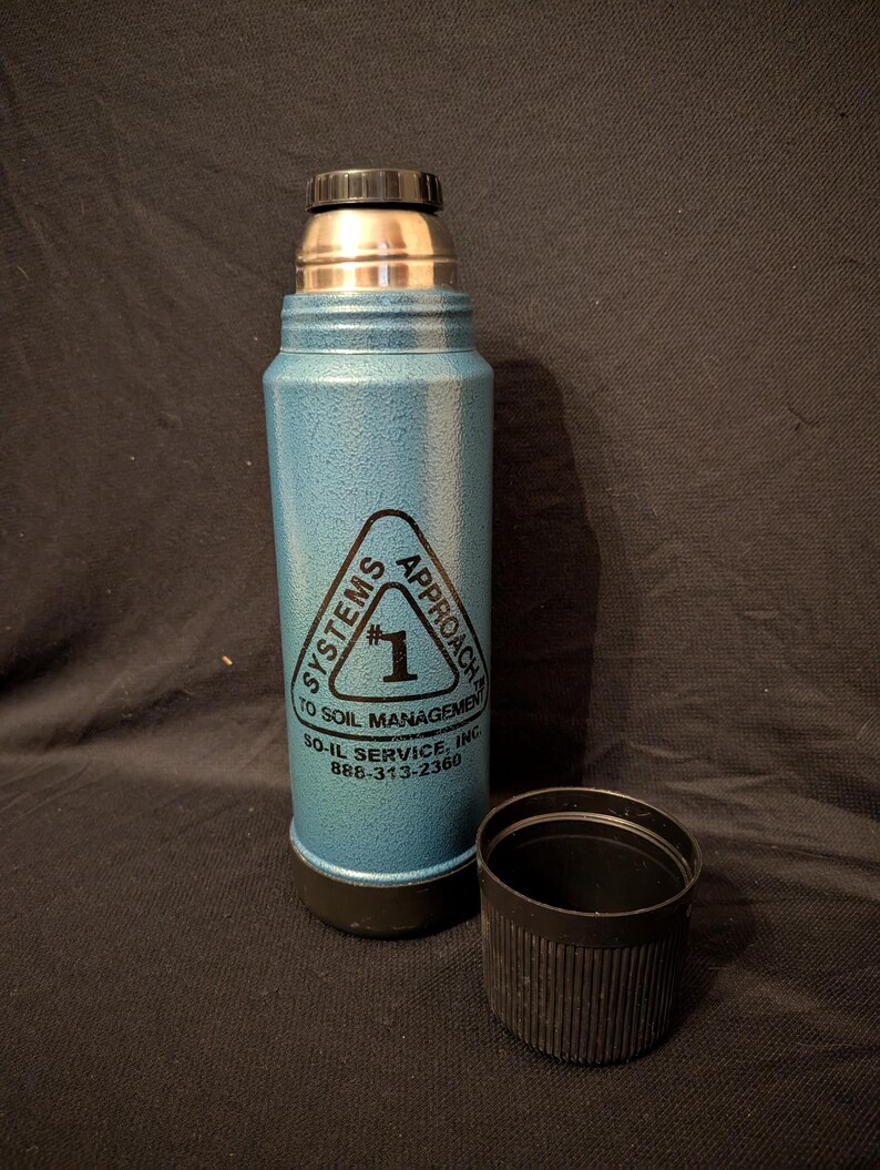 May include: A blue insulated thermos with a black lid and base, and a silver top. The thermos has a triangular logo with the text "SYSTEMS APPROACH TO SOIL MANAGEMENT" and the number "1". The black cap is next to the thermos.