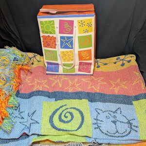 May include: A colorful gift bag with a white handle sits atop a woven blanket. The bag features a variety of square designs with suns, stars, and flowers. The blanket has a blue, pink, and yellow pattern with cat and star designs.
