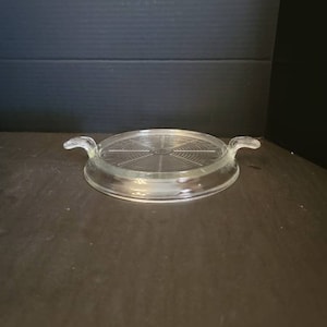 May include: A clear glass hot pad with a circular design and two handles. The hot pad is designed to protect surfaces from heat.
