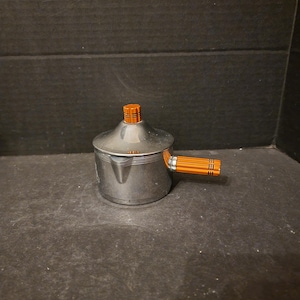 May include: A silver coffee pot with an orange handle and lid. The pot has a rounded body and a spout on the side.