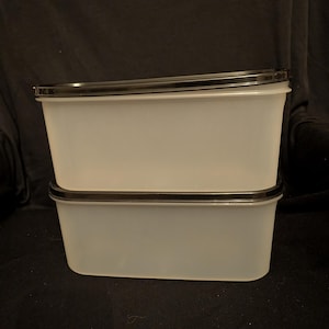 May include: Two rectangular, translucent food storage containers stacked on top of each other. Each container has a black rim around the top. The containers are made of a light, frosted plastic.