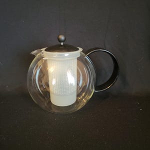 May include: A clear glass teapot with a black handle and a white mesh infuser. The teapot has a black lid with a small black knob on top.