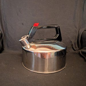 May include: A stainless steel tea kettle with a black handle and a red plastic lever on top. The kettle has a spout and a shiny, reflective surface. The handle is rectangular with an open center.