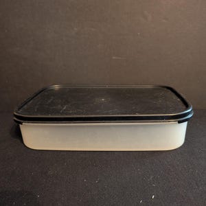 May include: A rectangular, translucent food storage container with a black lid. The container has a black rim and a slightly rounded base. The container is empty and sits on a dark surface.